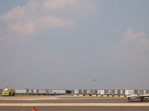 1988 Ford Bronco - A yellow tow vehicle is parked on an airport tarmac next to a line of trailers, with a plane flying in the distance.