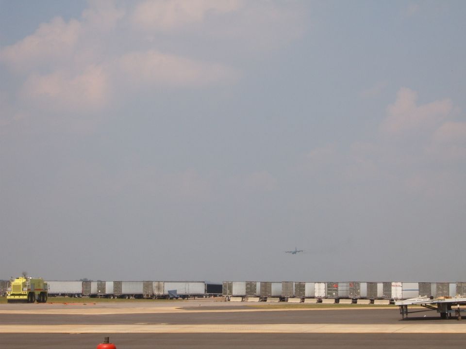 airport tarmac with a yellow tow vehicle, many parked trailers, and a plane in the sky