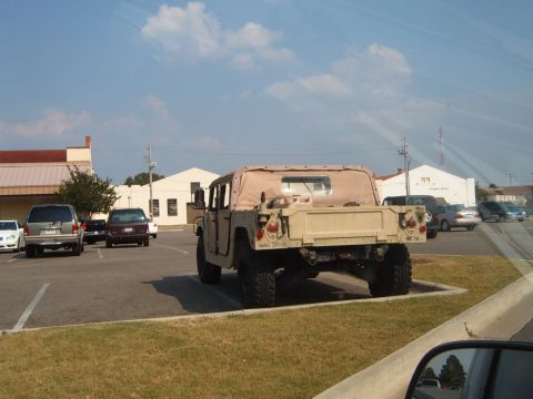 1988 Ford Bronco - A tan military vehicle is parked in a lot with other cars under a blue sky.
