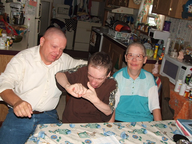 Three people sit at a table in a kitchen. One person in the middle is interacting with another's hands