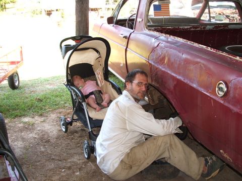 1966 Ford Ranchero - A man works on the wheel well of a rusty 1966 Ford Ranchero while a baby sleeps in a stroller beside him.