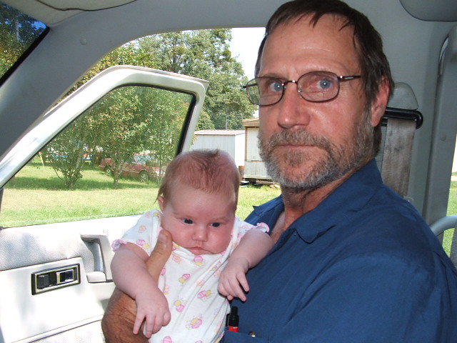 Man with glasses and beard holds a baby inside a vehicle; door is open, trees and buildings visible outside