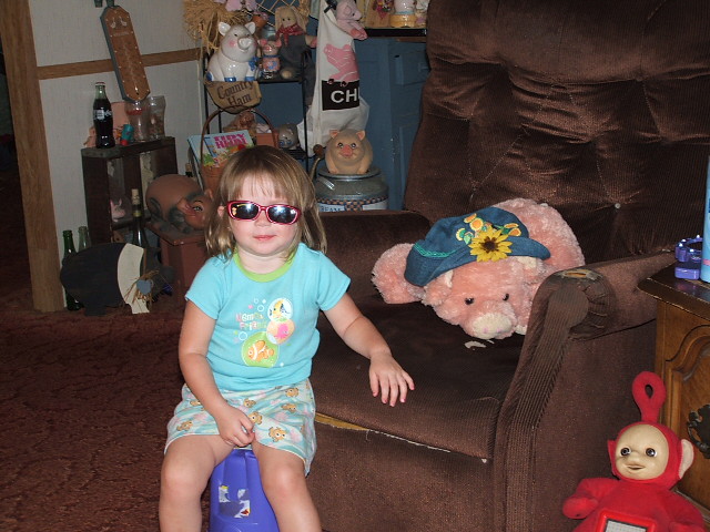 Young girl wearing sunglasses sits on a blue stool in a room with stuffed animals and furniture