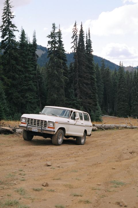 1979 Ford Bronco - This white Ford Bronco is parked on a dirt road surrounded by a forest and mountains.