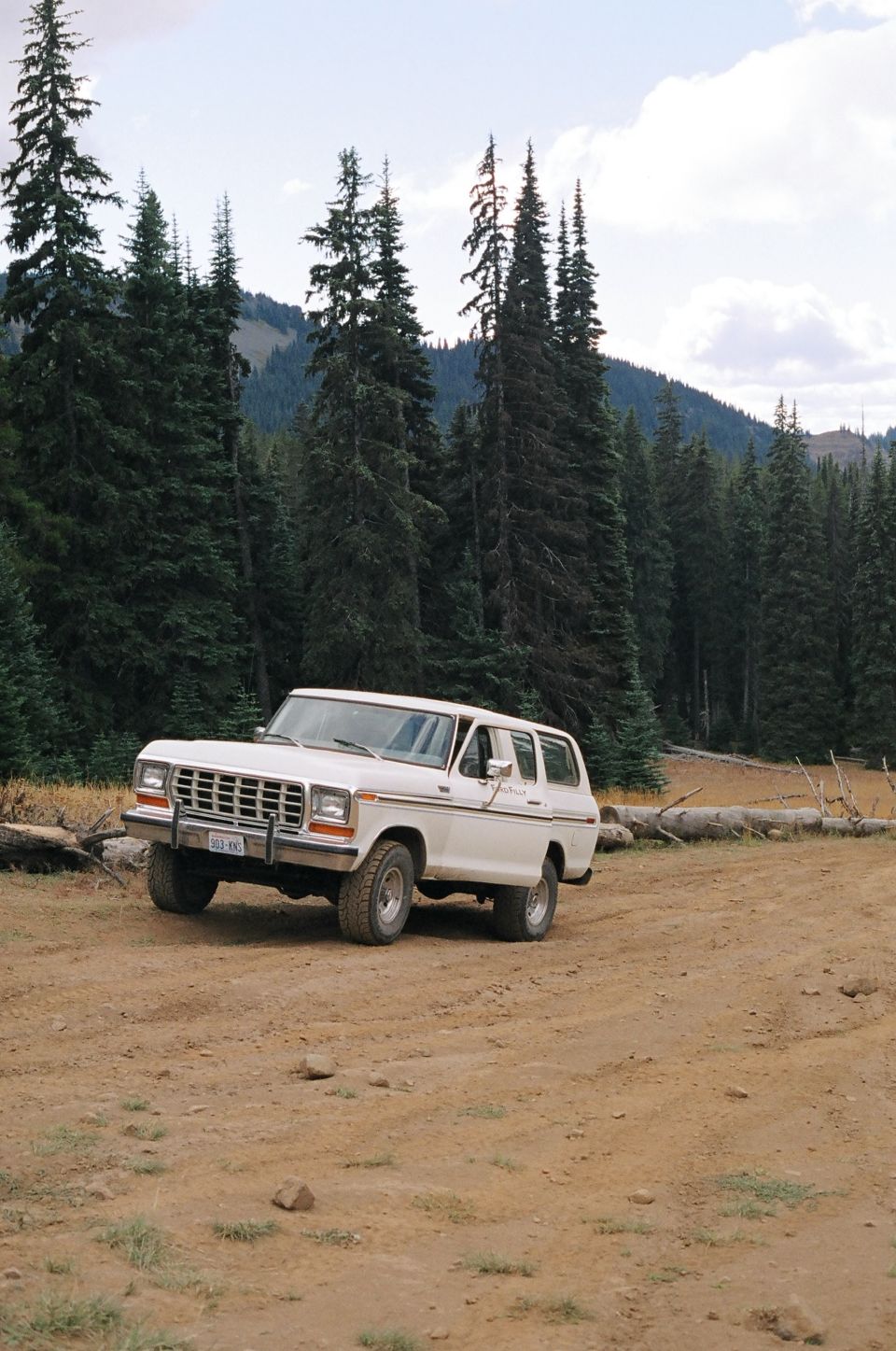 White 1978 Ford Bronco SUV parked on a dirt road with pine trees and mountains in the background