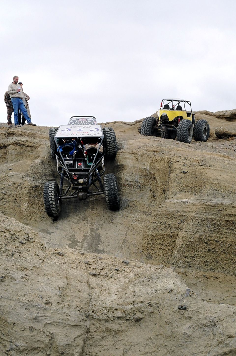 Two custom off-road vehicles navigate a steep, rocky terrain under an overcast sky