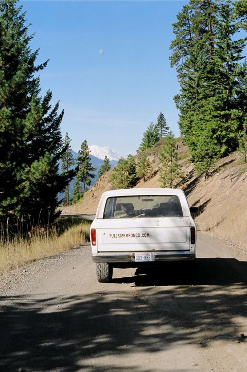 1979 Ford Bronco - A white Ford Bronco drives away on a dusty mountain road, with a snow-capped peak visible in the distance.