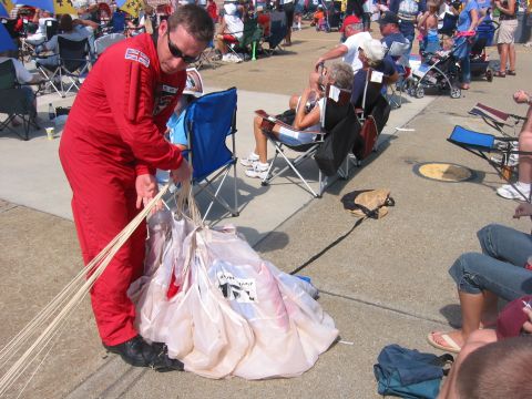 1989 Ford Bronco - A man in a red jumpsuit tends to a parachute on the ground, with an audience seated in the background at an air show.