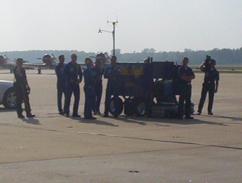 Navy-uniformed flight crew standing on an airfield beside a cart, jets parked in the background
