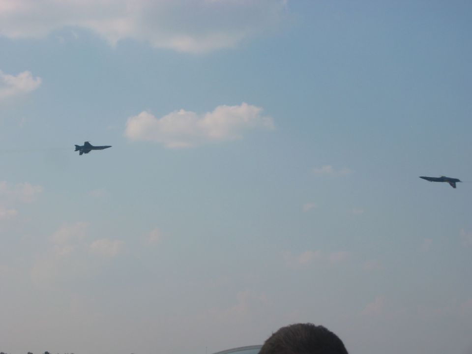 Two dark fighter jets in a pale blue sky with scattered clouds. One jet is on the left, the other on the right