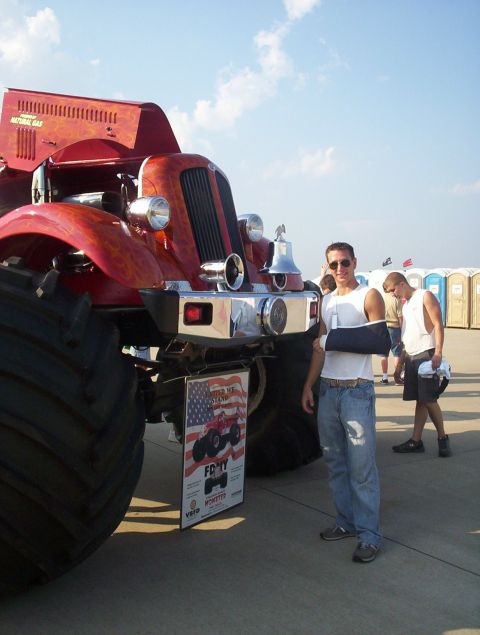 1989 Ford Bronco - A man with a cast on his arm stands next to a large red monster truck at an outdoor event.