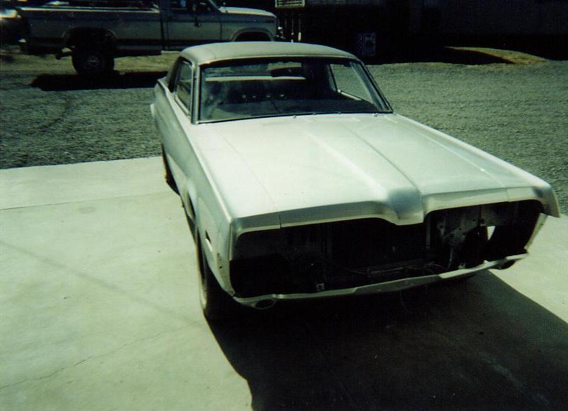 Silver coupe with missing grille and headlights, parked on concrete. A pickup truck is visible in the background
