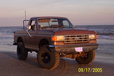 1987 Ford Bronco - A lifted Ford pickup truck sits on a sandy beach with the ocean in the background at sunset.