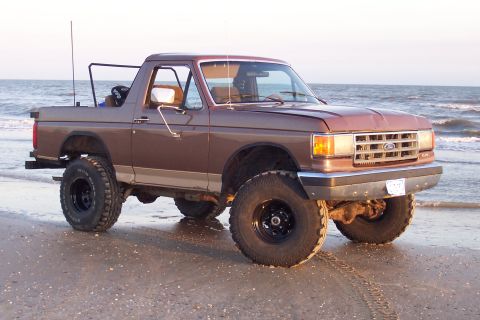 1987 Ford Bronco - Brown two-tone Ford Bronco with its fiberglass top removed, parked on wet sand at the water's edge with surf breaking behind it. It is lift…