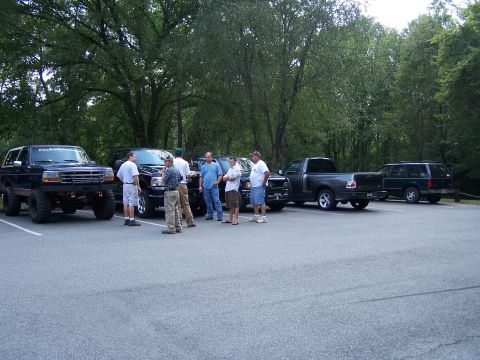 1979 Ford F-150 - A group of men gather around several trucks and SUVs in a parking lot surrounded by trees.