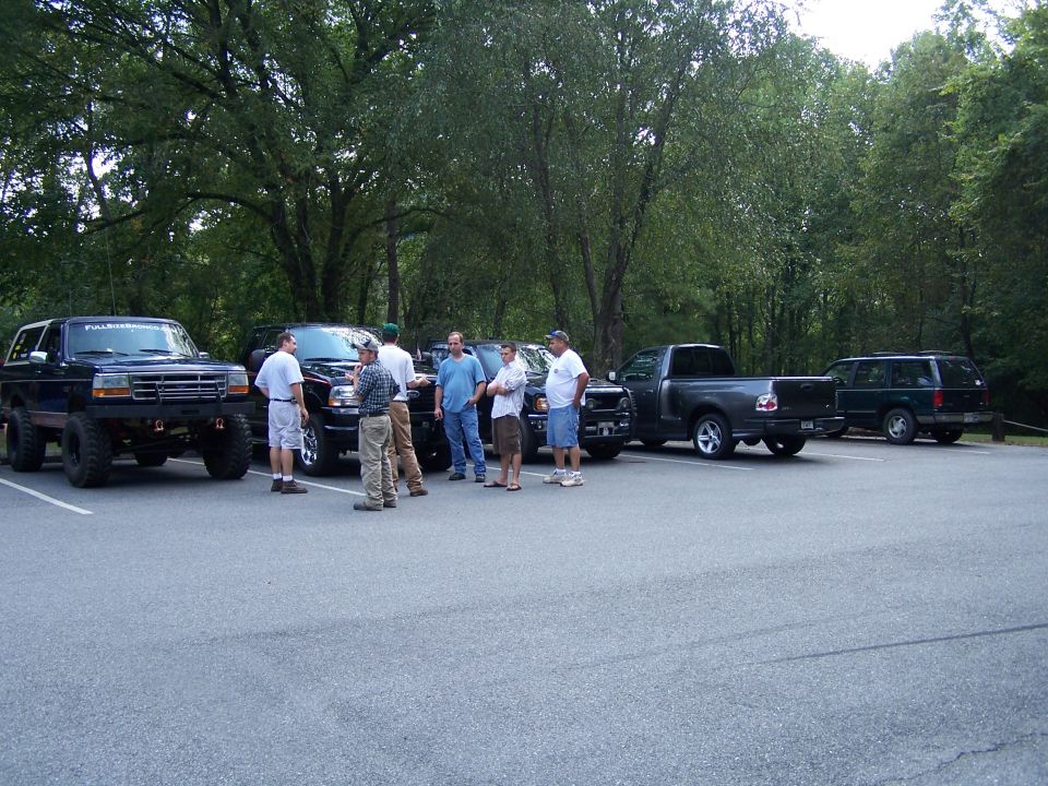 Several guys stand around a collection of trucks and SUVs parked in a lot near trees