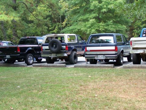 1979 Ford F-150 - Several trucks and SUVs are parked in a grassy lot with trees in the background.