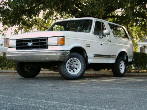 1988 Ford Bronco - This white Ford Bronco is parked on a street, with trees overhead and a house in the background.