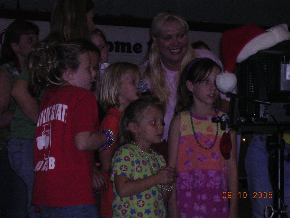 Children and adults gathered, some wearing necklaces, with a Santa hat on a monitor