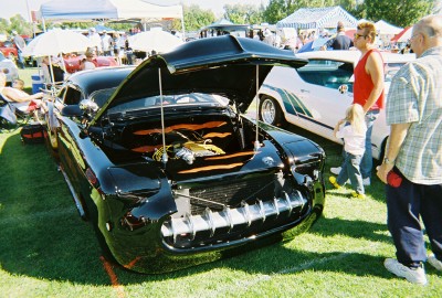1972 Ford Ranchero - A custom black vehicle with its hood raised is displayed on a grassy field, with spectators in the background.