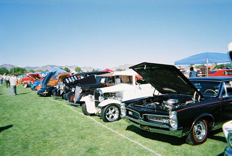 Multiple classic cars lined up on a grassy field, hoods open, under a clear blue sky