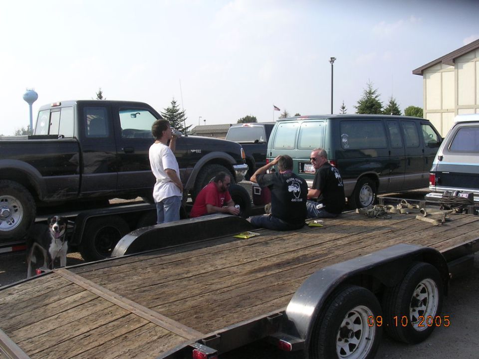 people sitting on a trailer next to a black pickup truck and a dark green van