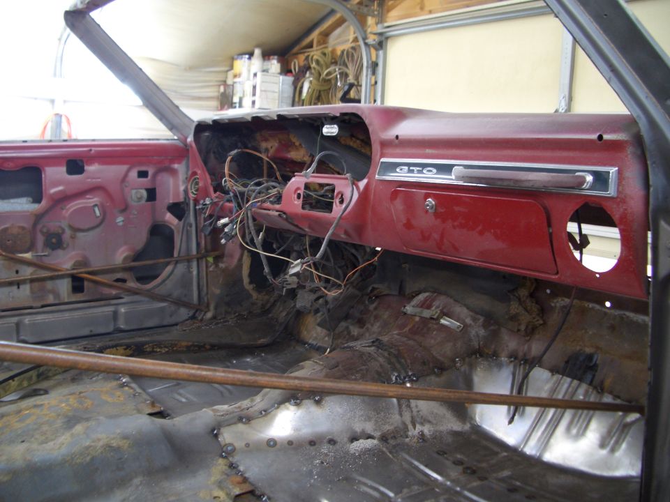 Stripped red GTO interior with exposed wiring, GTO dash badge, and bare metal floors during restoration