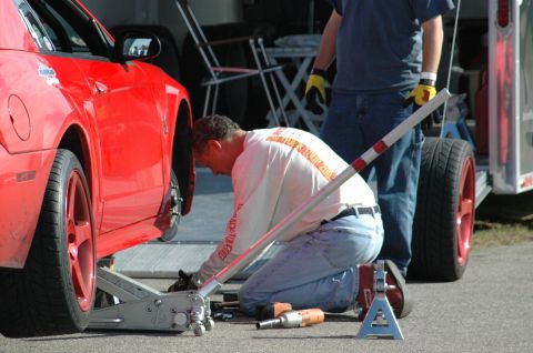 2000 Ford Mustang - A mechanic works on a red sports car, which is lifted by a jack, with a trailer and spare tire in the background.