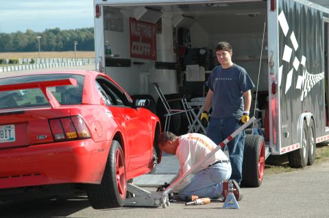 2000 Ford Mustang - A red Ford Mustang is being serviced next to a trailer, with two people present at what appears to be a race track or event.