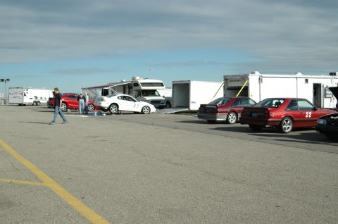 2000 Ford Mustang - Multiple vehicles, including a red coupe and a white car, are parked in a lot with trailers under a cloudy sky.