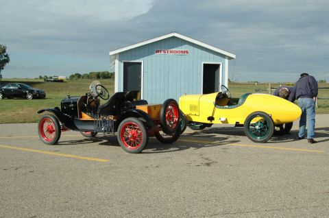 2000 Ford Mustang - Two vintage race cars, one black and one yellow, are parked side-by-side in front of a blue restroom building.