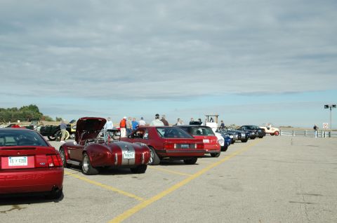 2000 Ford Mustang - A collection of vehicles, including a modern red Mustang and a classic red convertible, are parked in a lot with spectators.