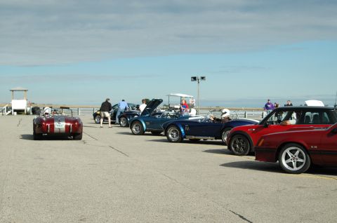 2000 Ford Mustang - A group of vintage-style roadsters are parked on a racetrack, with people milling around.