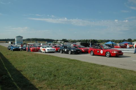 2000 Ford Mustang - A group of cars are staged on a race track under a clear blue sky with scattered clouds.