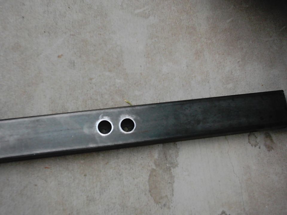 Rectangular metal tube with two circular holes drilled near one end, resting on a concrete surface