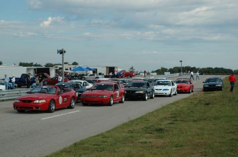 2000 Ford Mustang - A group of Ford Mustangs are gathered on a paved track under a cloudy sky, with trailers and tents in the background.