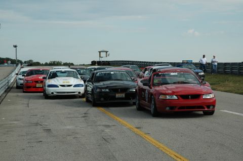 2000 Ford Mustang - Multiple Ford Mustangs line up on a race track, with the closest vehicle being a red model.
