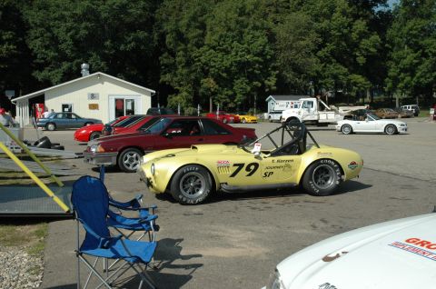 2000 Ford Mustang - A yellow race car with racing decals is parked in a lot with other vehicles and a building.
