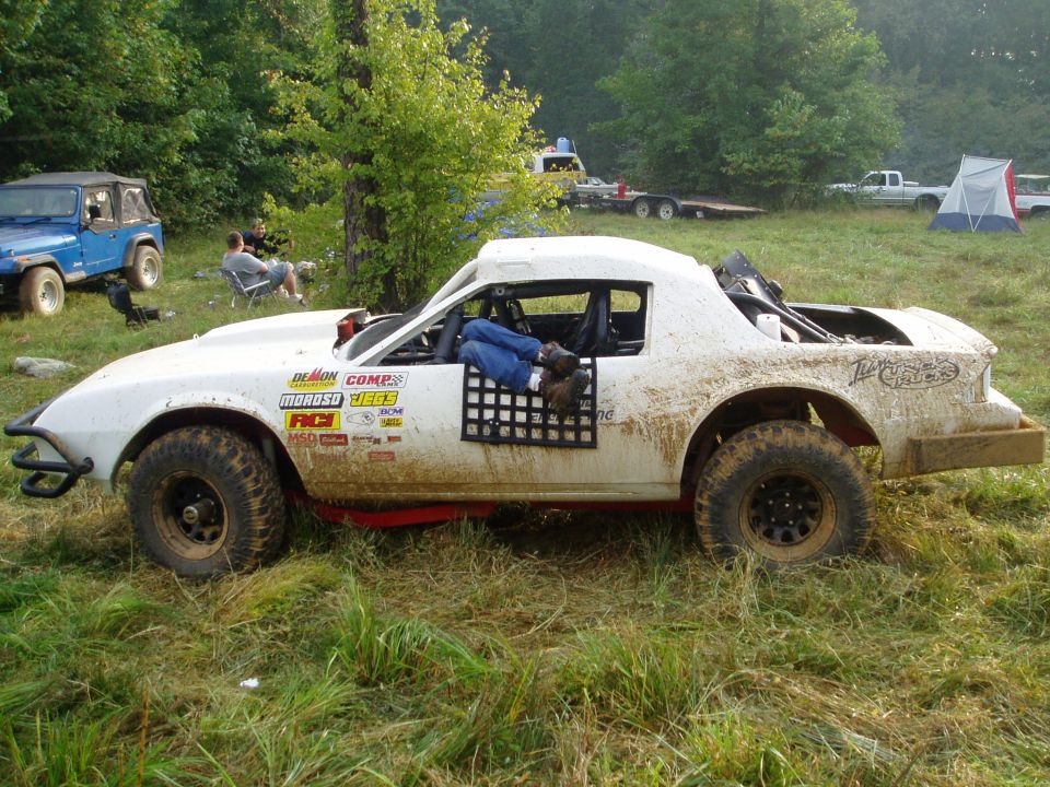 Muddy white caged off-road race coupe on knobby tires in a grassy pit area