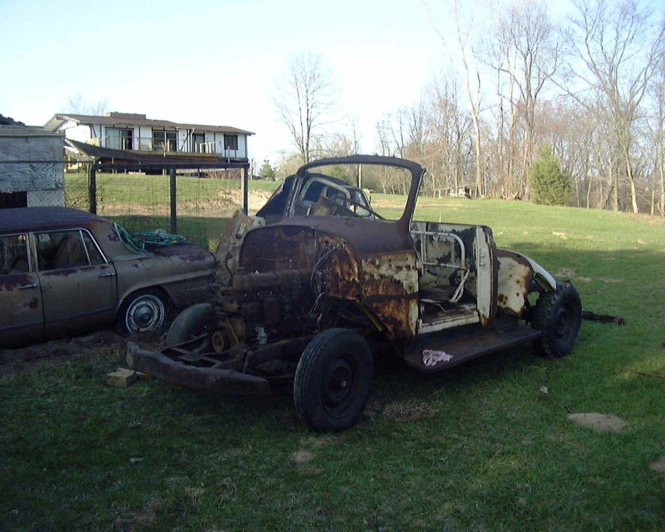 Rusted stripped vehicle body on frame in grassy yard beside old sedan