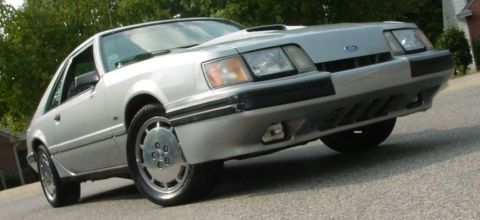 1986 Ford Mustang - A silver coupe is shown from a low front quarter angle, parked outdoors on a paved surface.