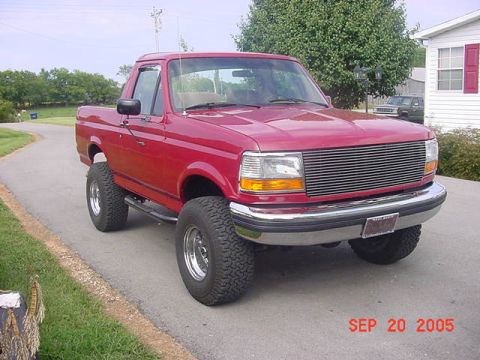 1989 Ford Bronco - Three-quarter front view of a lifted red full-size Ford Bronco with the hardtop removed, parked in a residential driveway beside a white ho…