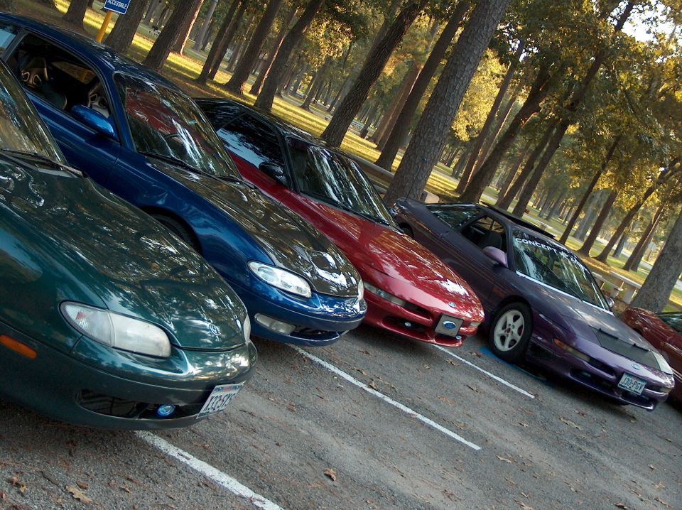 Several Ford Probes parked in a line on a sunny day with trees in the background. One is green, one is blue, one is red, and one is purple