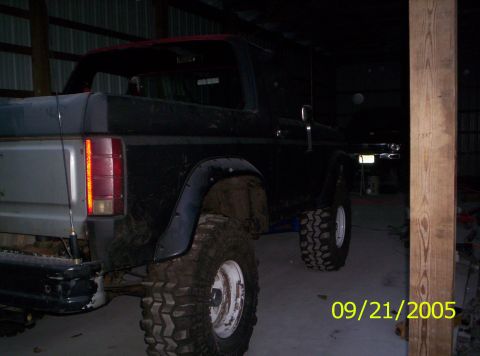 1985 Ford Bronco - This lifted Ford Bronco, seen from the rear, features large off-road tires and a dark body with a red roof.