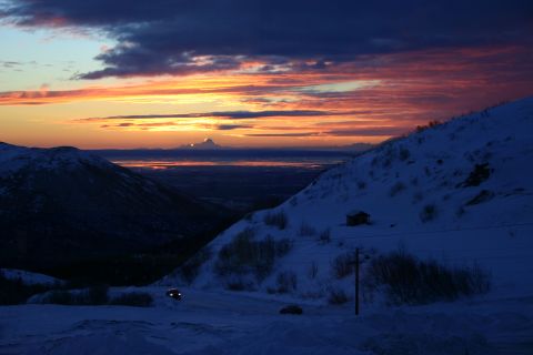 2003 Jeep TJ - A scenic view of a snow-covered mountain range during a vibrant sunset, with a distant cabin and vehicles on a road.