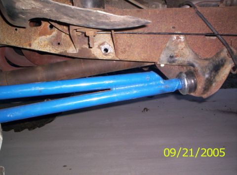 1985 Ford Bronco - Close-up of a blue suspension arm connected to a rusty vehicle frame, with exhaust pipes underneath.