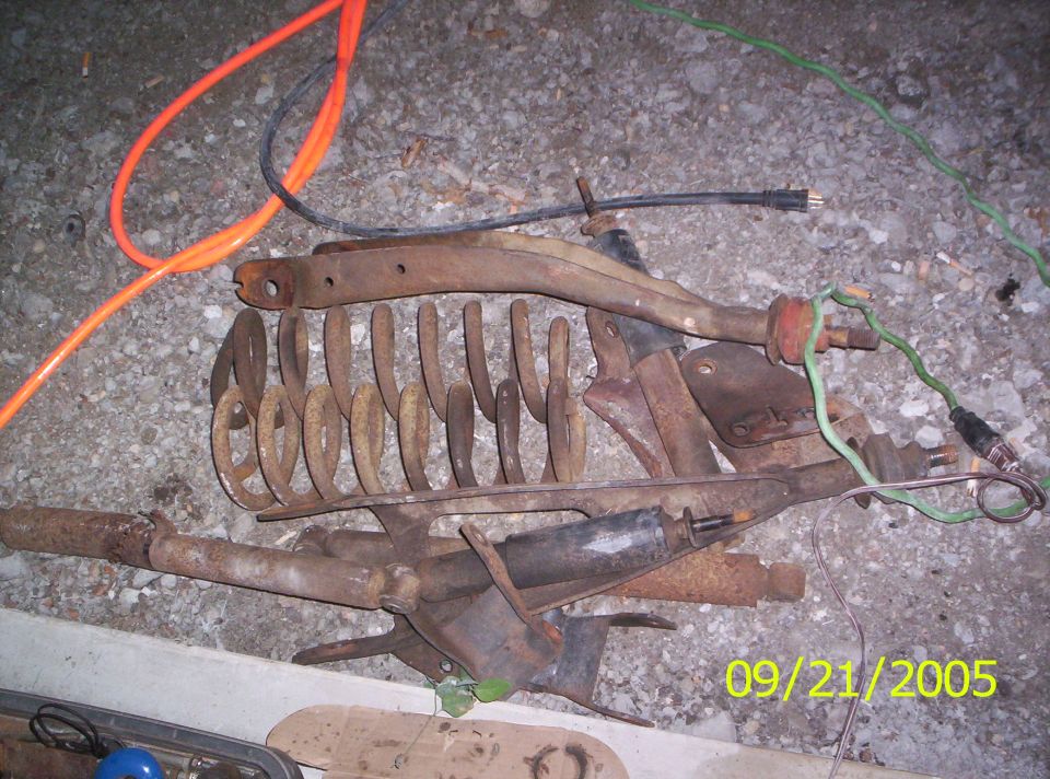 Pile of rusty suspension parts, including springs and control arms, with orange and black cords on the ground
