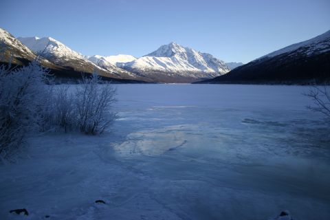 2003 Jeep TJ - A vast, frozen lake stretches towards snow-capped mountains under a bright blue sky, with frosted bushes in the foreground.