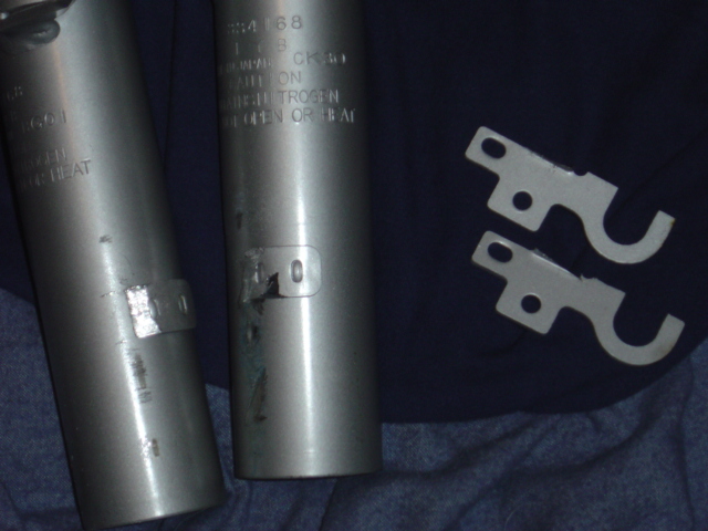 Two metal cylinders with markings and two metal brackets on a dark fabric surface