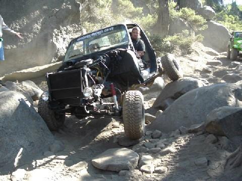 1996 Ford Bronco - An off-road vehicle navigates a rocky trail, its hood open, with another green vehicle visible in the background.